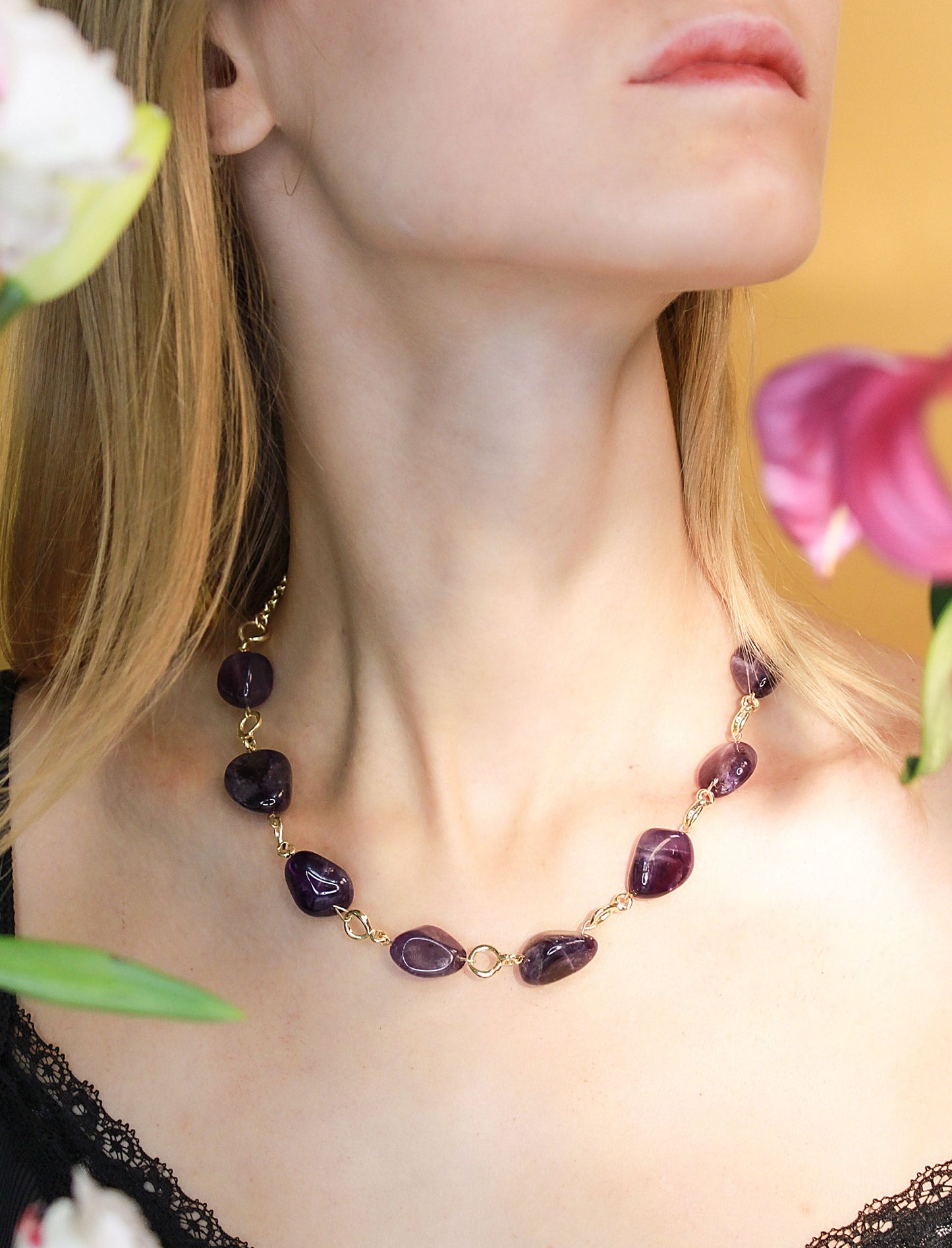 Close-up of a necklace with purple gemstones worn by a person, with flowers in the background.