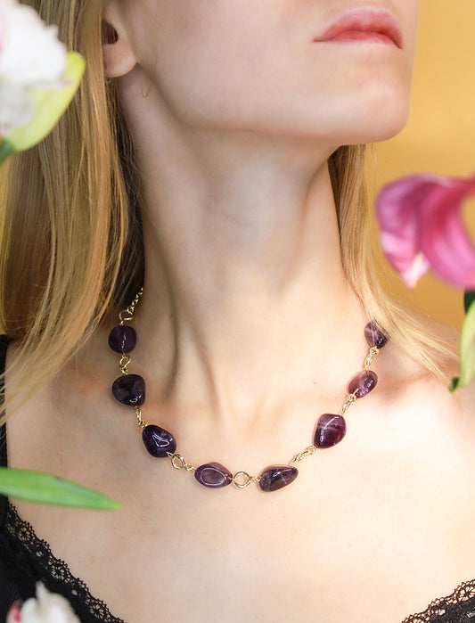 Close-up of a necklace with purple gemstones worn by a person, with flowers in the background.